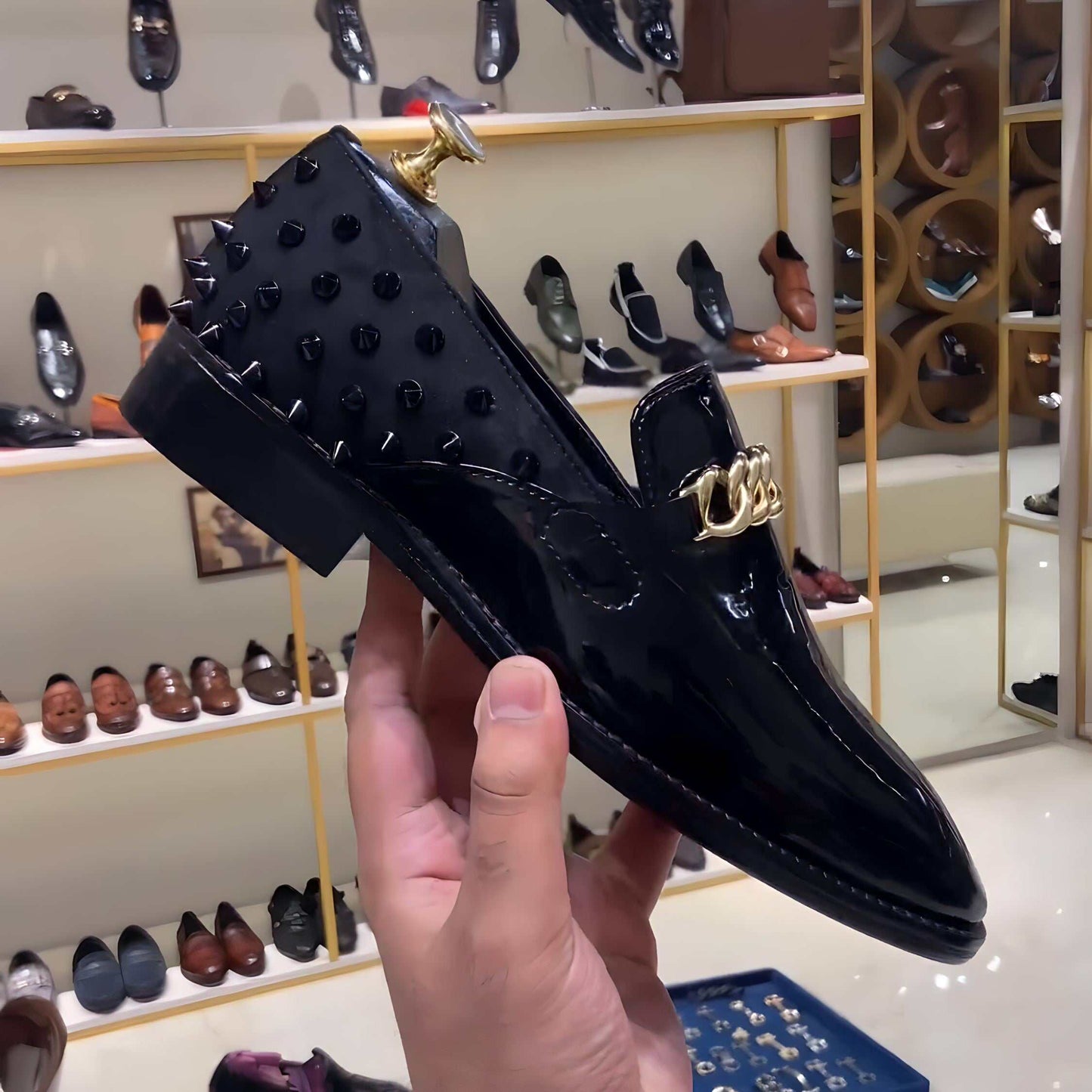 Black studded patent moccasins with gold chain detail held in hand in shoe store.